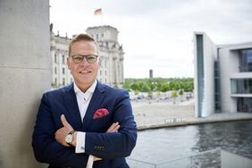 Ein Mann im Anzug mit Einstecktuch und Brille blickt freundlich in die Kamera. Im Hintergrund ist der Deutsche Bundestag zu sehen.